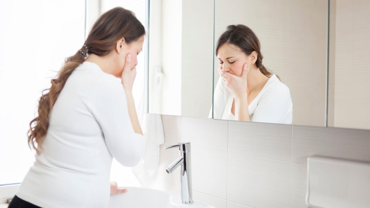  Pregnant woman pictured standing in front of a bathroom mirror holding her mouth as if she's going to be ill. 