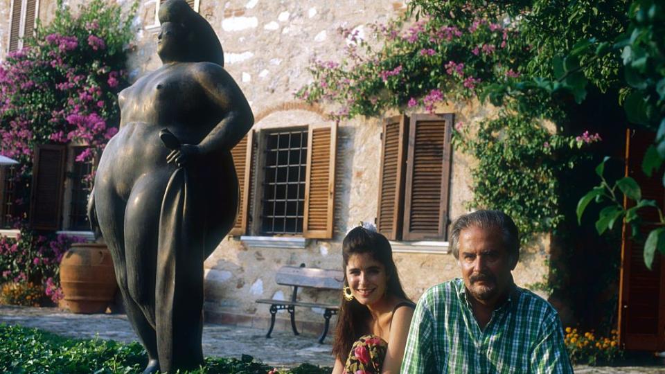 sophia vari and fernando botero sit on the ground and pose for a photo in a backyard next to a large bronze sculpture