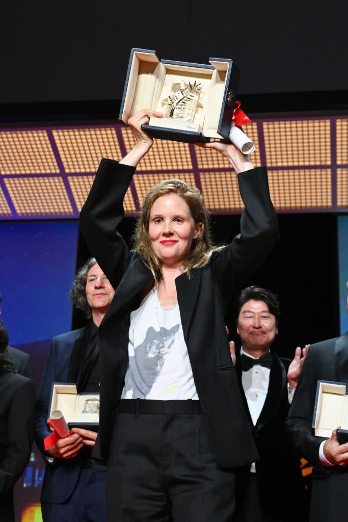 justine triet holds an open box above her head with both hands, she wears a black suit with a white shirt, people stand behind her