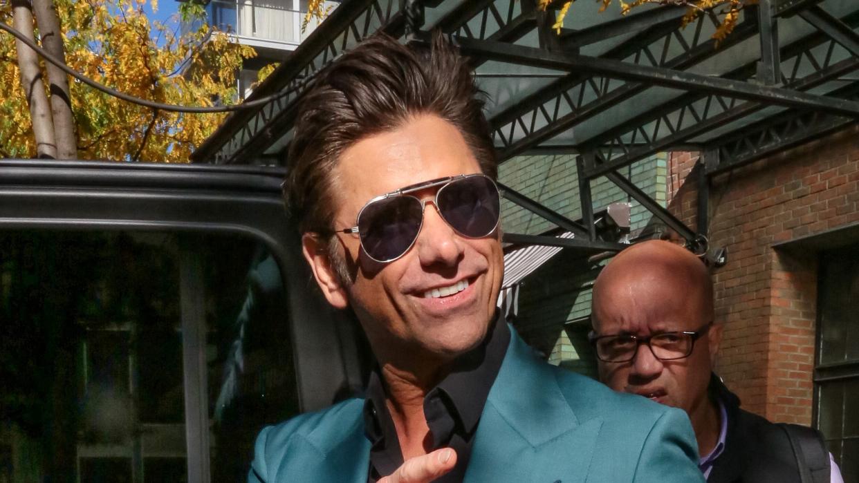 john stamos smiles and points at someone off camera, he wears a teal suit jacket and black collared shirt with aviator sunglasses and holds a copy of his memoir if you would have told me