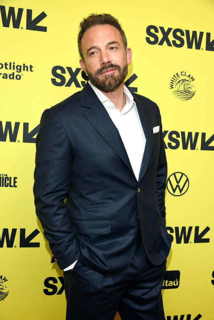 Ben Affleck stands on the SXSW event red carpet, wearing a tailored dark suit and white dress shirt, hands in pockets, with sponsor logos in the background