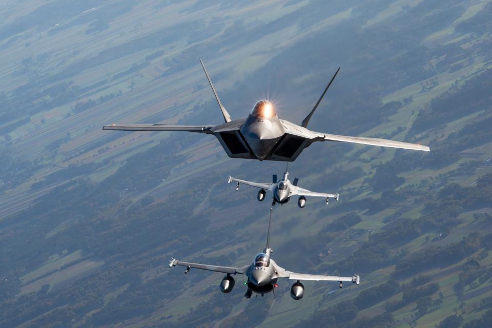 A US Air Force F-22 Raptor flies above two Polish F-16 jets against a lush and green background.