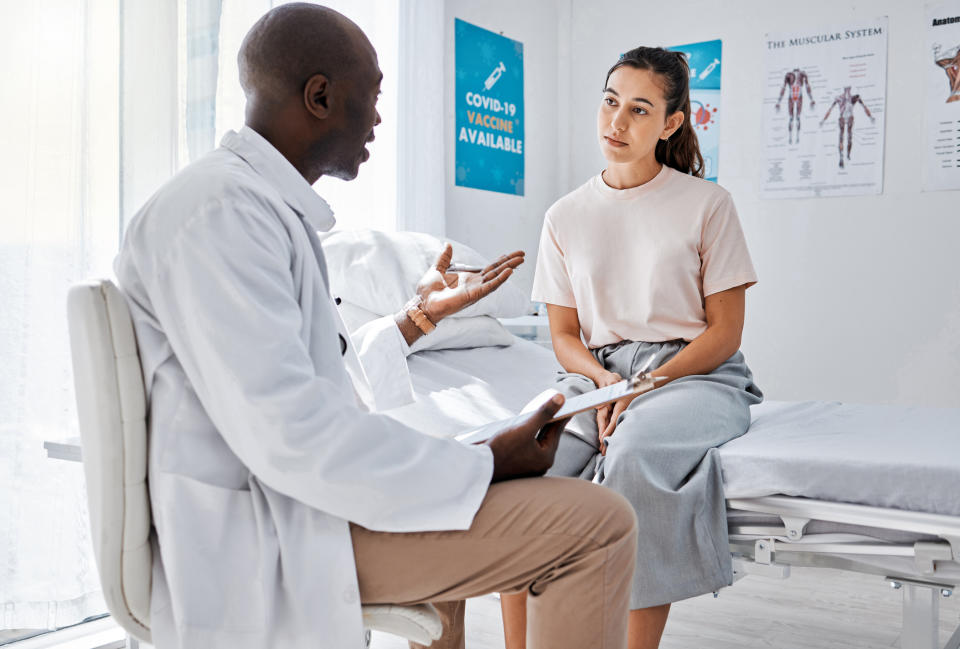 A doctor speaks with a seated patient in a medical office. Posters about vaccination and the muscular system are on the wall behind them