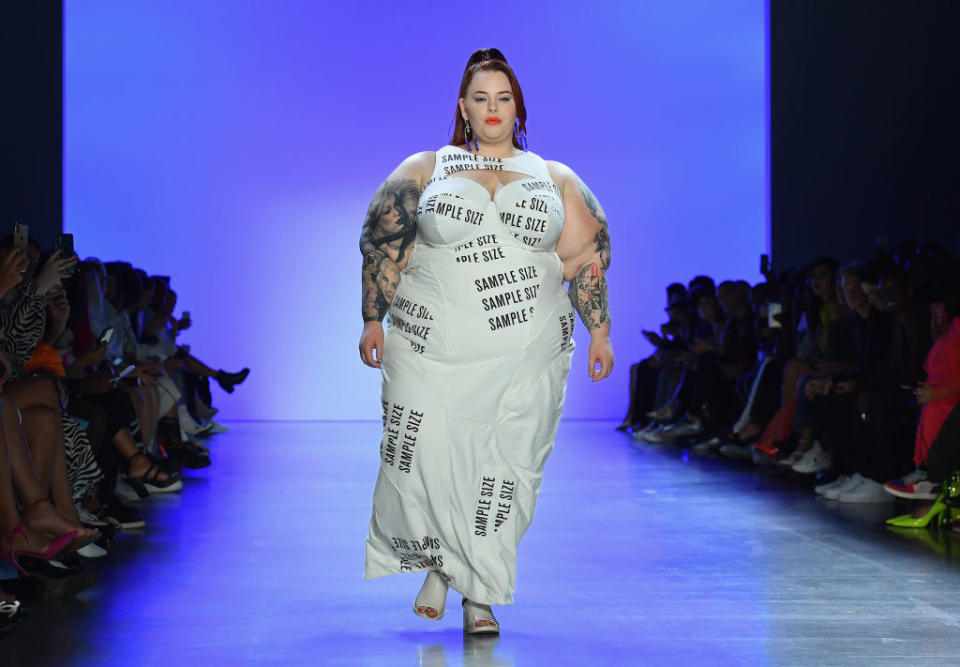 Tess Holliday walking the catwalk wearing a full white dress with "Sample size" printed all over it