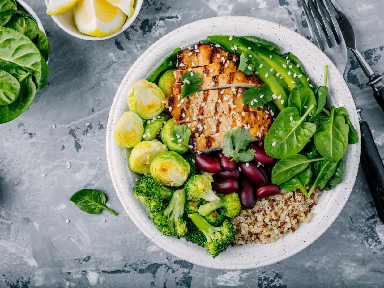 Healthy buddha bowl lunch with grilled chicken, quinoa, spinach, avocado, brussels sprouts, broccoli, red beans with sesame seeds on dark gray background. Top view.