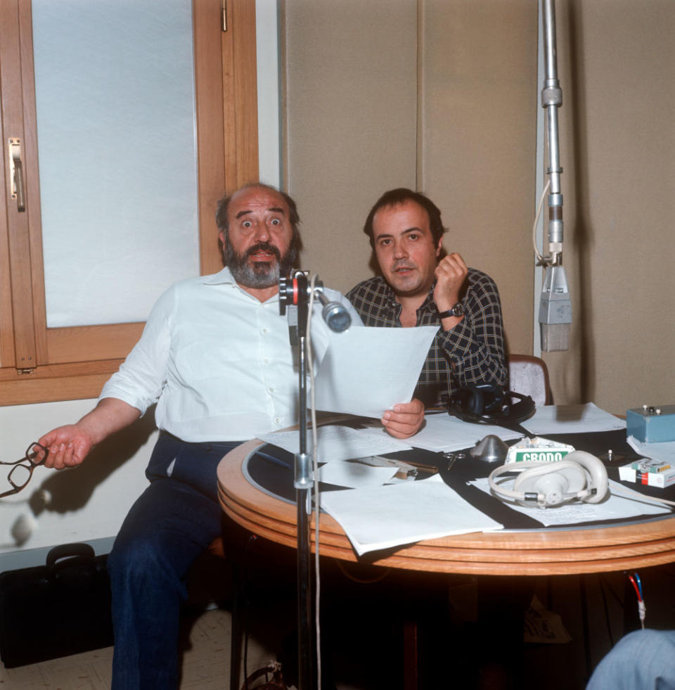 Two men sit at a round table with audio equipment and papers. One man holds eyeglasses and a sheet of paper; the other rests his chin on his hand