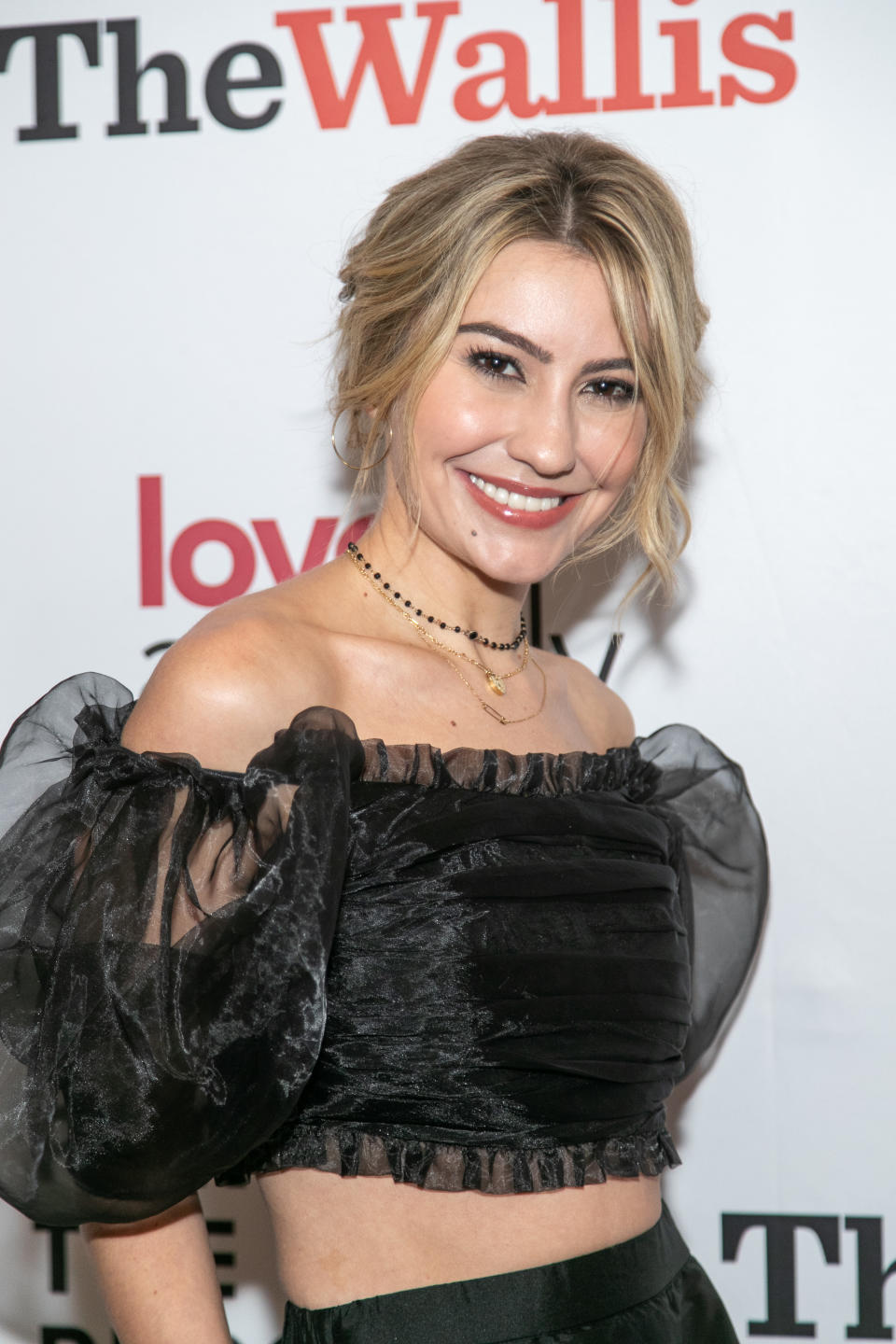 Woman in an off-the-shoulder black top with sheer sleeves at a red carpet event