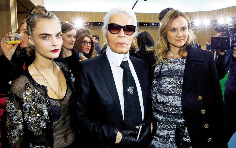 The designer, surrounded by photographers and standing between the two actresses, wears his signature white pony tail and dark glasses