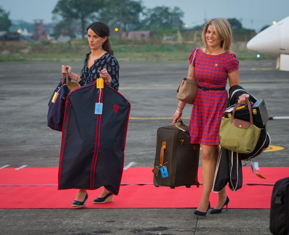 Two women in heels and dresses holding suitcases and garment bags.