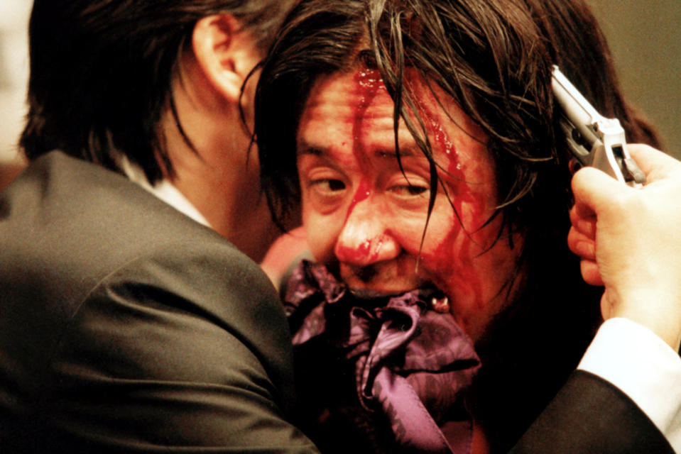 Min-sik's character covered in blood while another character puts a gun to his head