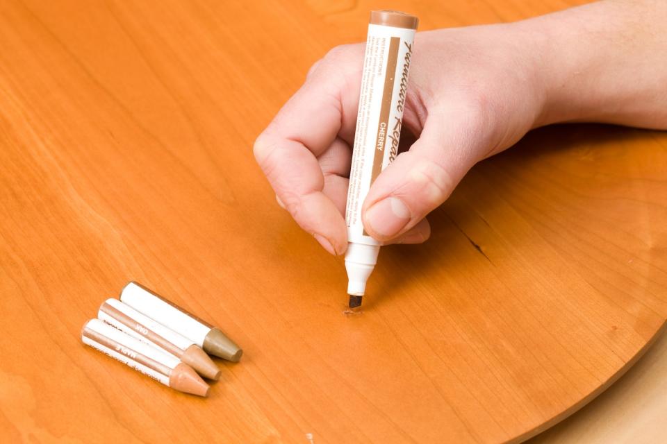 A worker is using a furniture repair marker to color fix a scratched cherry table surface. Three repair sticks rest to the left.  ( How to remove permanent marker from plastic)