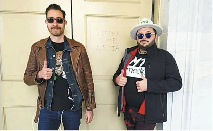 Two men posing with thumbs up outside a beige door that has 