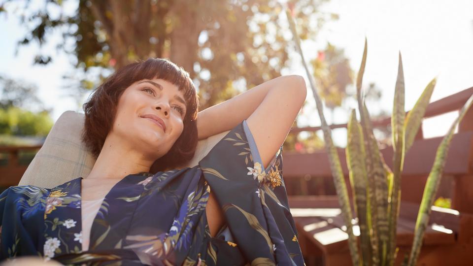 Young woman looking deep in thought and smiling while lying back in a deck chair on her patio on a sunny afternoon.