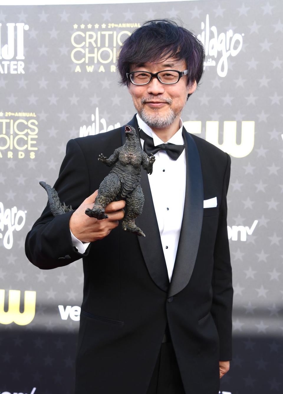 takashi yamazaki smiles at the camera and holds a godzilla figurine in front of him, he wears a black tuxedo and glasses