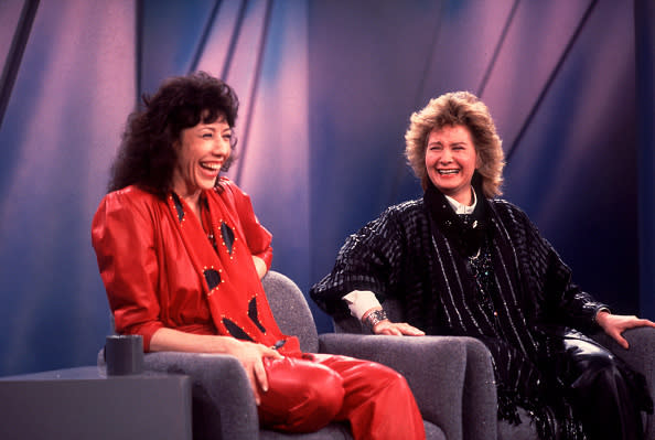 Lily Tomlin and Linda Bloodworth-Thomason seated on a talk show stage, both smiling and laughing. Lily wears a red outfit, Linda wears a black sequined outfit