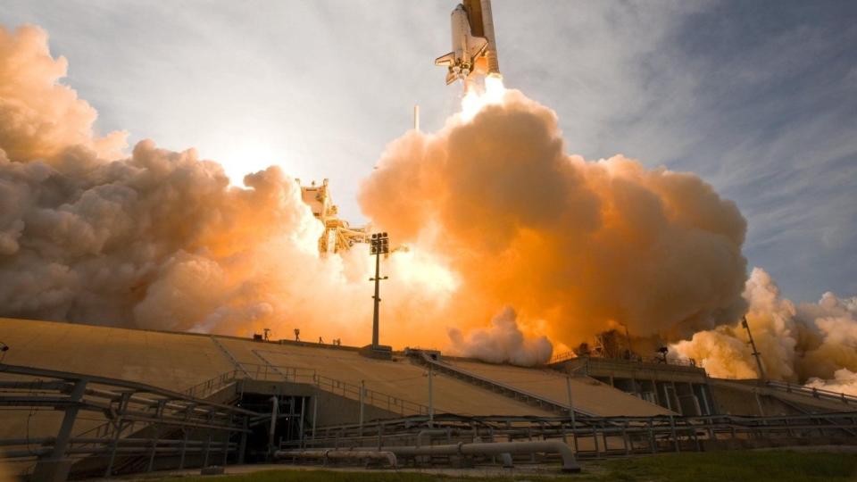 Ein Spaceshuttle startet mit viele Feuer und Qualm von der Erde.