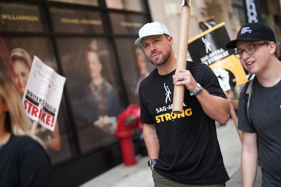 Stephen Amell holds a sign while walking the SAG-AFTRA picket line in New York. He wears a white baseball cap and a "SAG-AFTRA strong" black t-shirt.