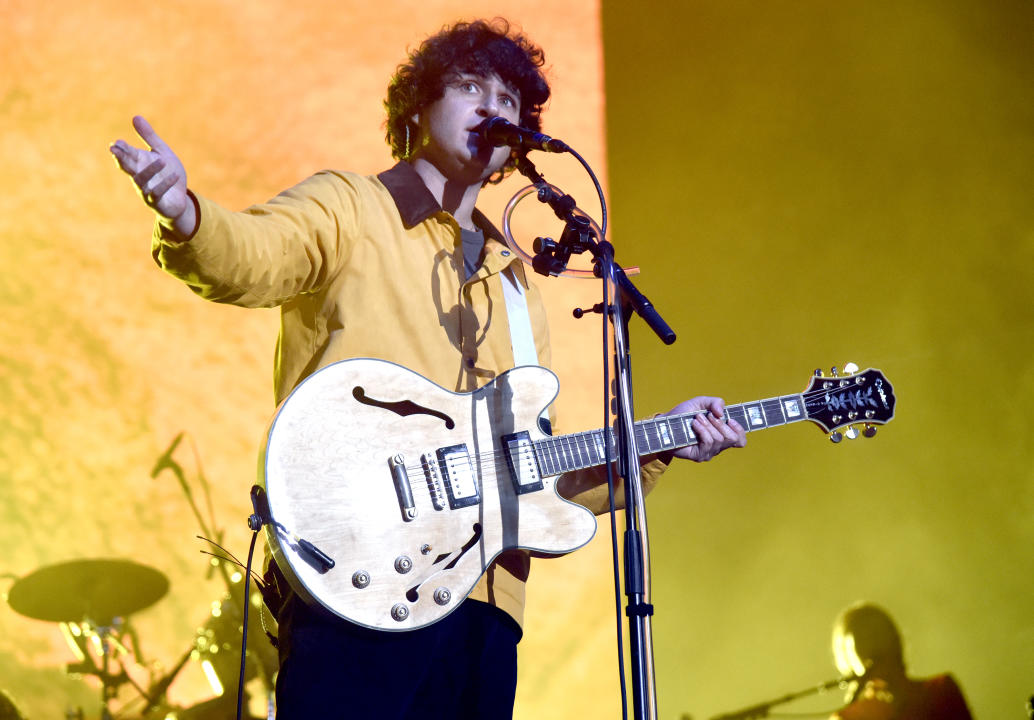 Ezra Koenig of Vampire Weekend stands at a microphone while holding a guitar.