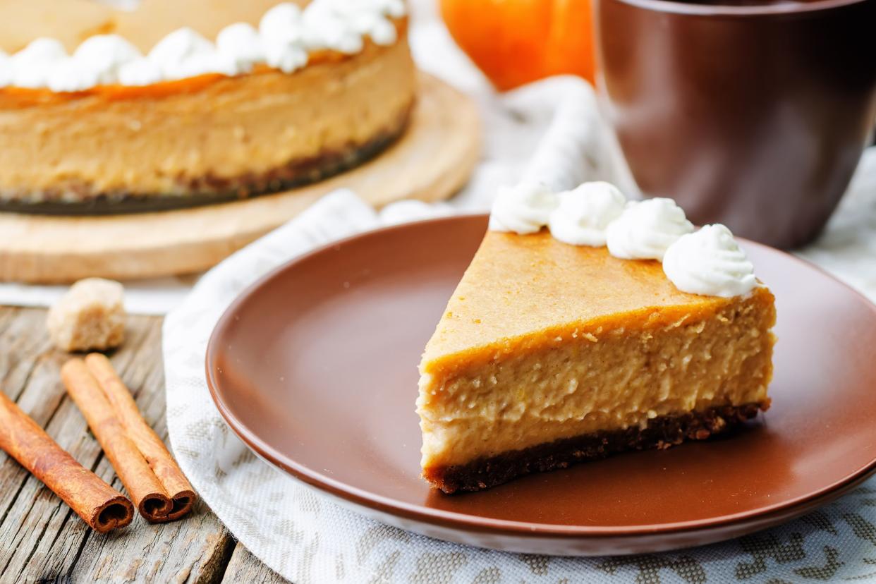 A piece of pumpkin-maple crustless cheesecake on a rust colored plate with the rest of the cheesecake, cinnamon sticks, a brown coffee cup, and a pumpkin in the background