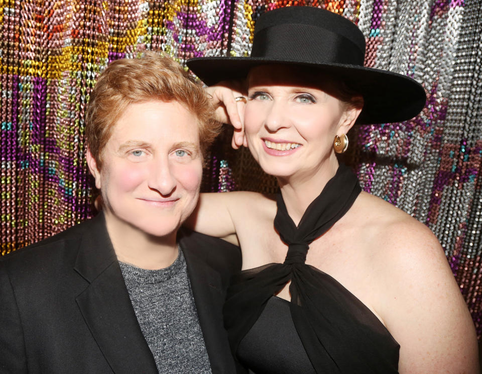 Cynthia Nixon and Christine Marinoni pose together, smiling, with Cynthia in a black dress and hat, and Christine in a blazer and top