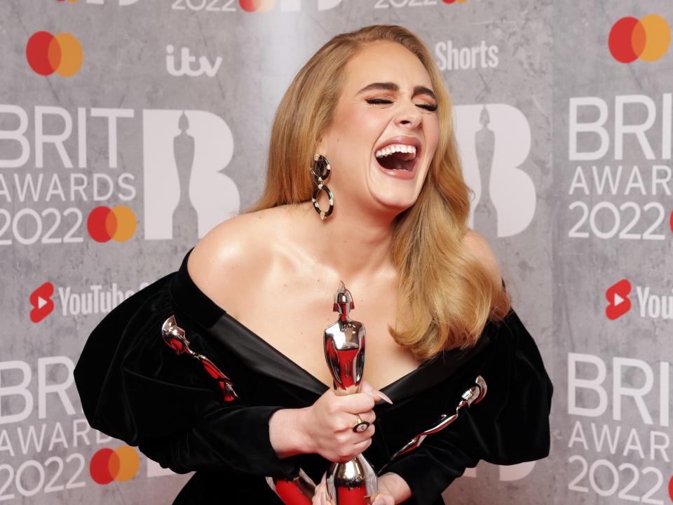 Adele laughing with her mouth wide open and eyes shut. She's leaning over and holding 3 awards.
