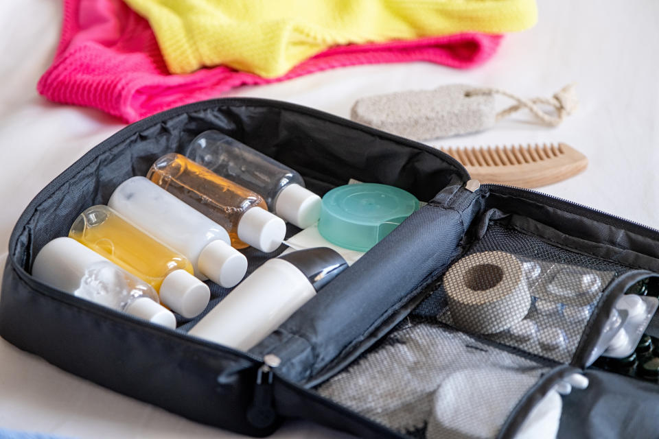 An open toiletry bag containing various travel-sized bottles, toiletries, a comb, and a pumice stone, with folded clothes in the background