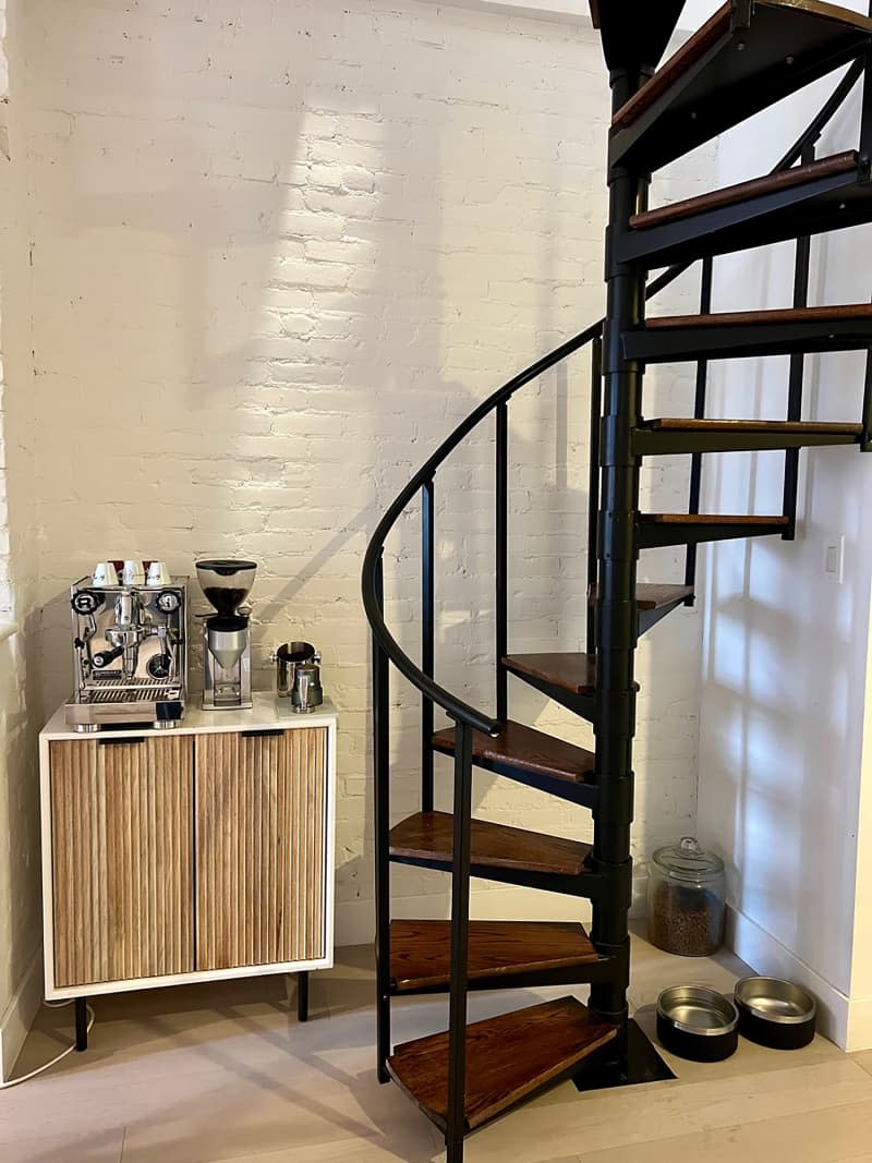 Coffee maker and grinder on side cupboard against painted brick wall and pet food and bowls flank black and brown wood spiral staircase.