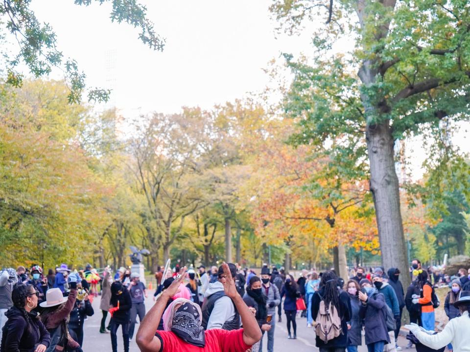 A crowd in Central Park surrounded by trees with red, orange, and green leaves.