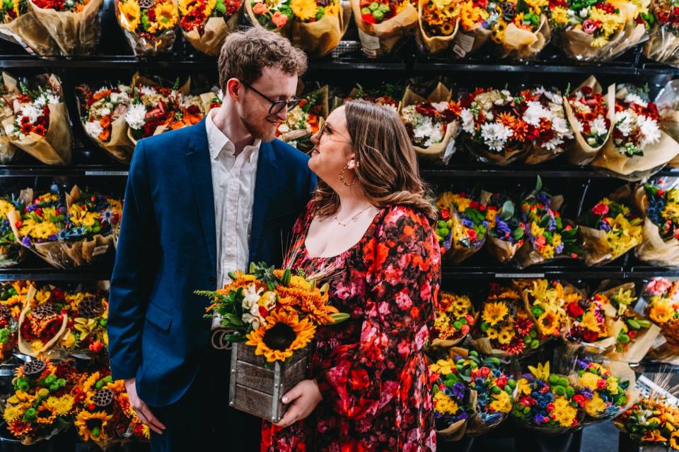 A couple look at each other in front of a display of flowers while holding a bouquet.