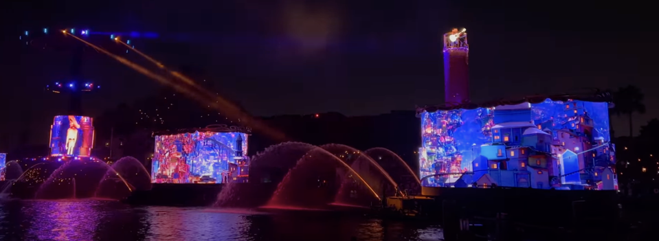 Nighttime water show with illuminated projections on floating screens and a water fountain display. Multiple colors and patterns light up the dark sky in the background