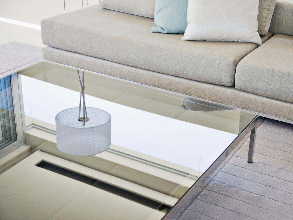 A glass-top table reflecting a lamp with a white shade in front of a gray couch on top of a gray carpet