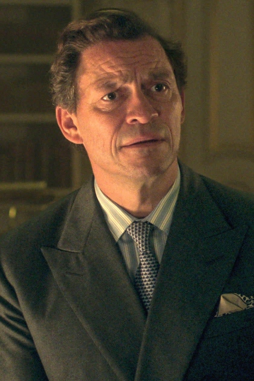 Dominic West, wearing a double-breasted suit, sits and listens attentively to a conversation with another character from The Crown. Bookshelves are in the background