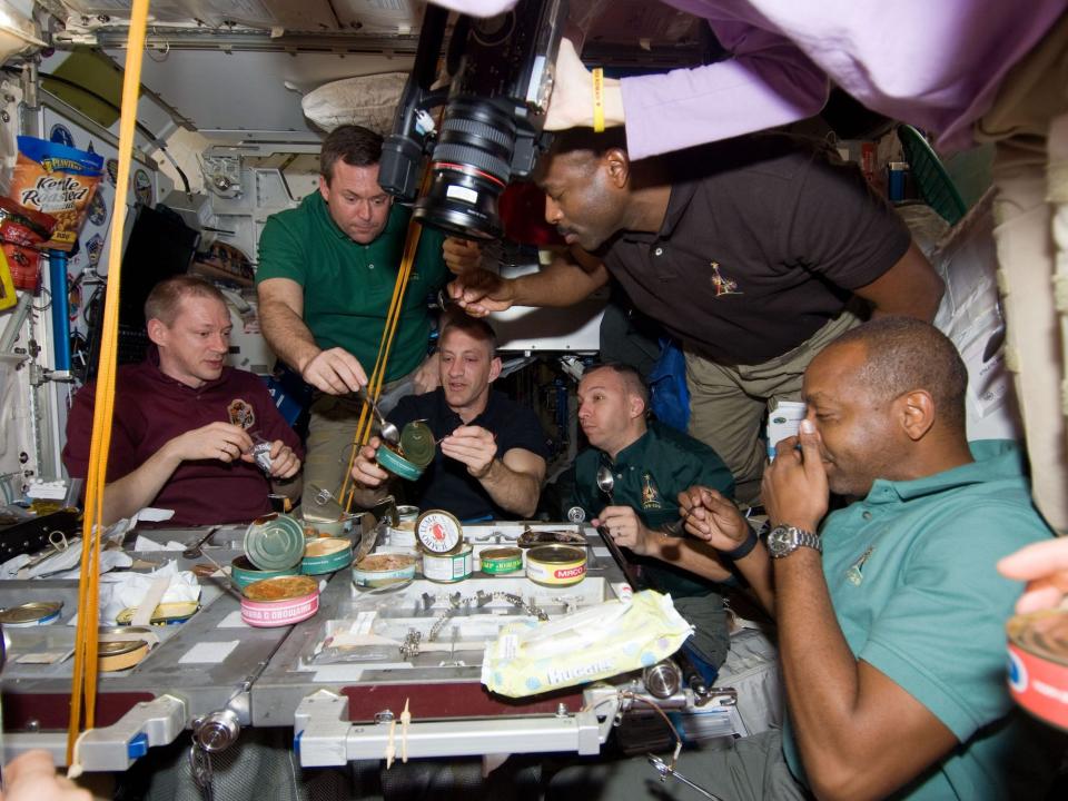 Several people gather around a table with cans and other food on it at the International Space Station
