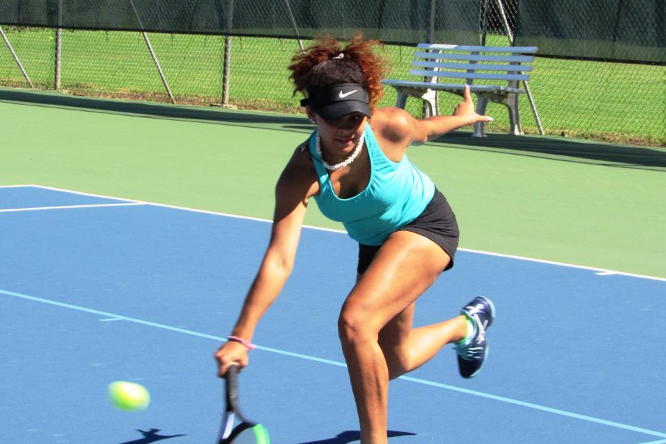 A high school girl wearing a blue tank top and black shorts hitting a ball with a tennis racket on a blue tennis field.