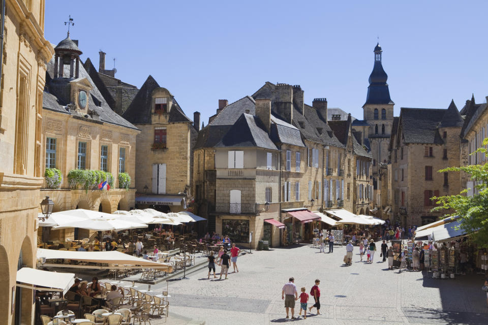 Historic village square with bustling market, people browsing stalls and terraced cafes under sunny sky