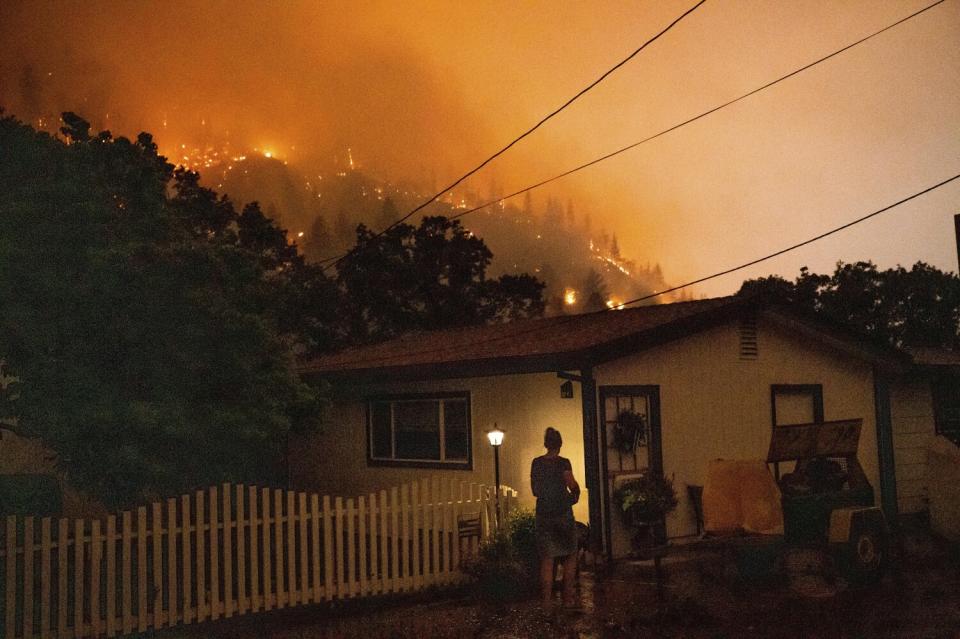 A woman outside a house framed by a burning ridge nearby.