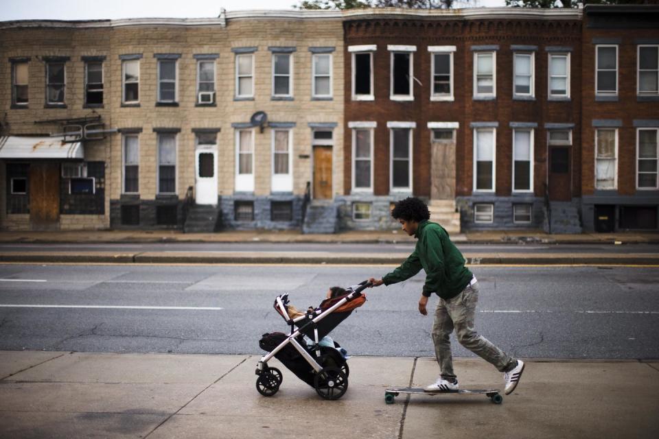 A young Black man on a skateboard pushes his son in a stroller on a sidewalk past blighted buildings in Baltimore.