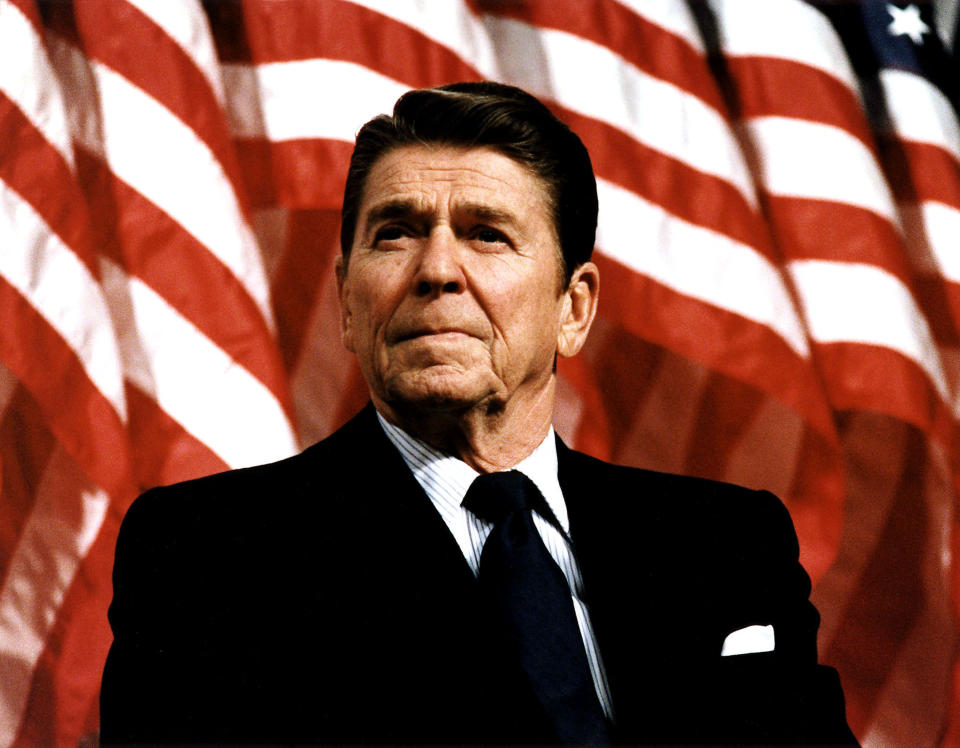 Ronald Reagan is standing in front of multiple American flags, dressed in a dark suit, white dress shirt, and tie