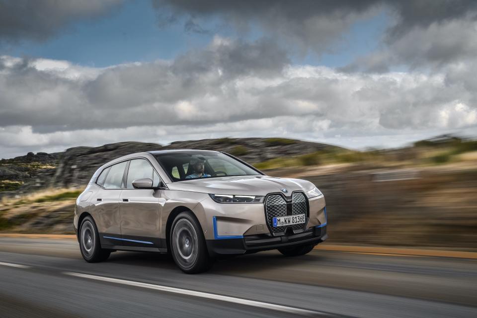 A silver BMW iX driving on a rural road against a cloudy sky.