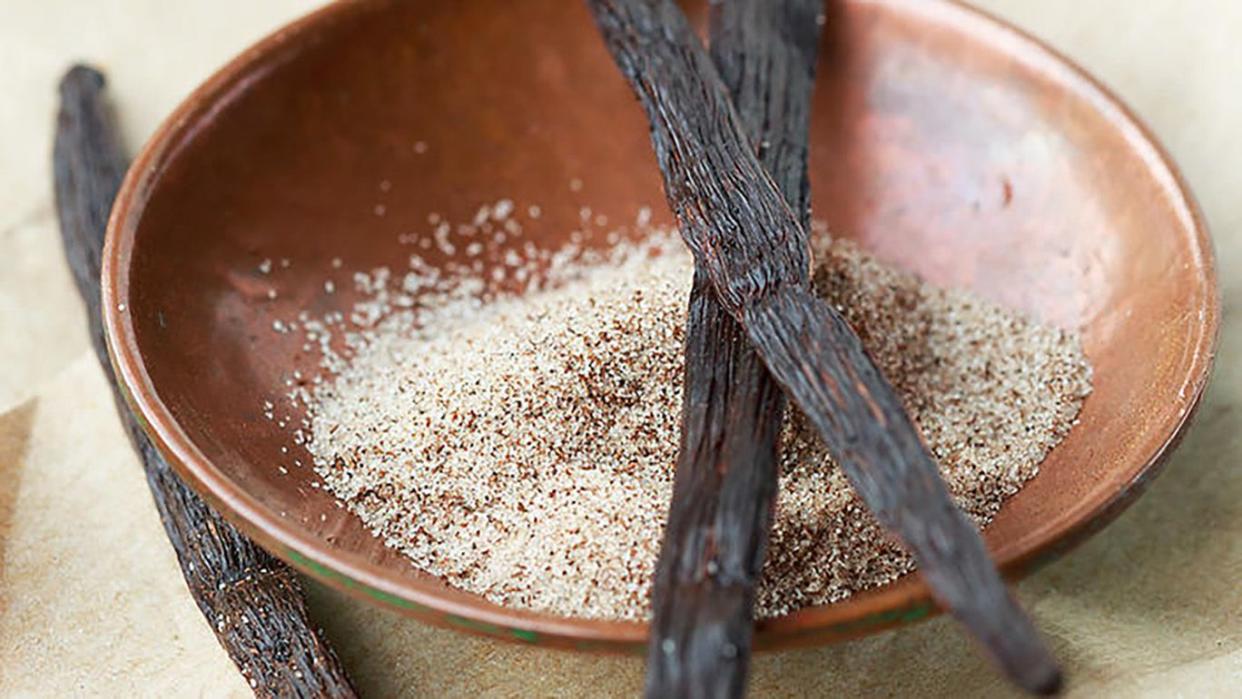 Two dried vanilla pods placed across a wooden bowl filled with sugar, and another dried vanilla pod next to the bowl.