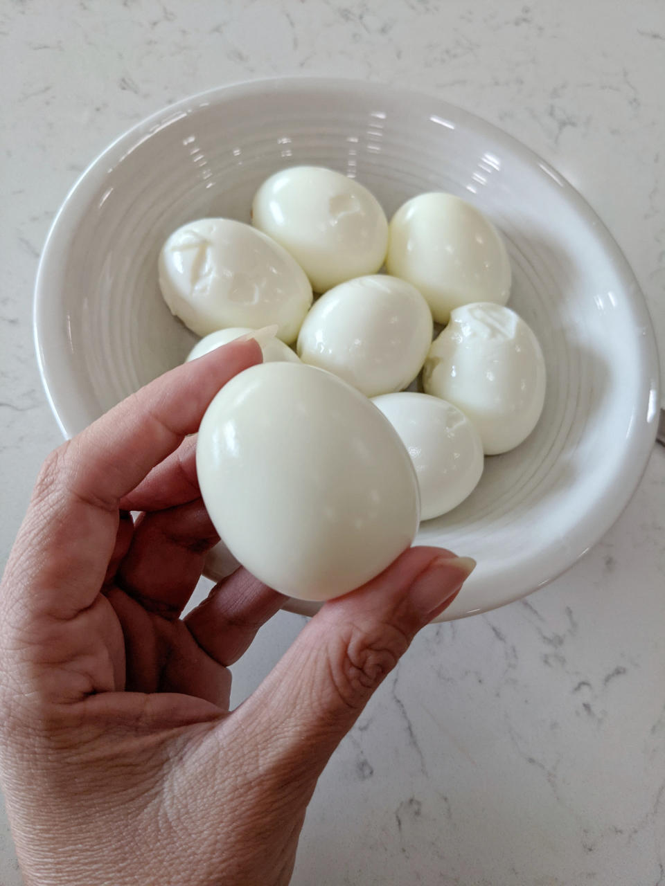 Woman's hand holding a peeled Hard-boiled egg in front of bowl of eggs.