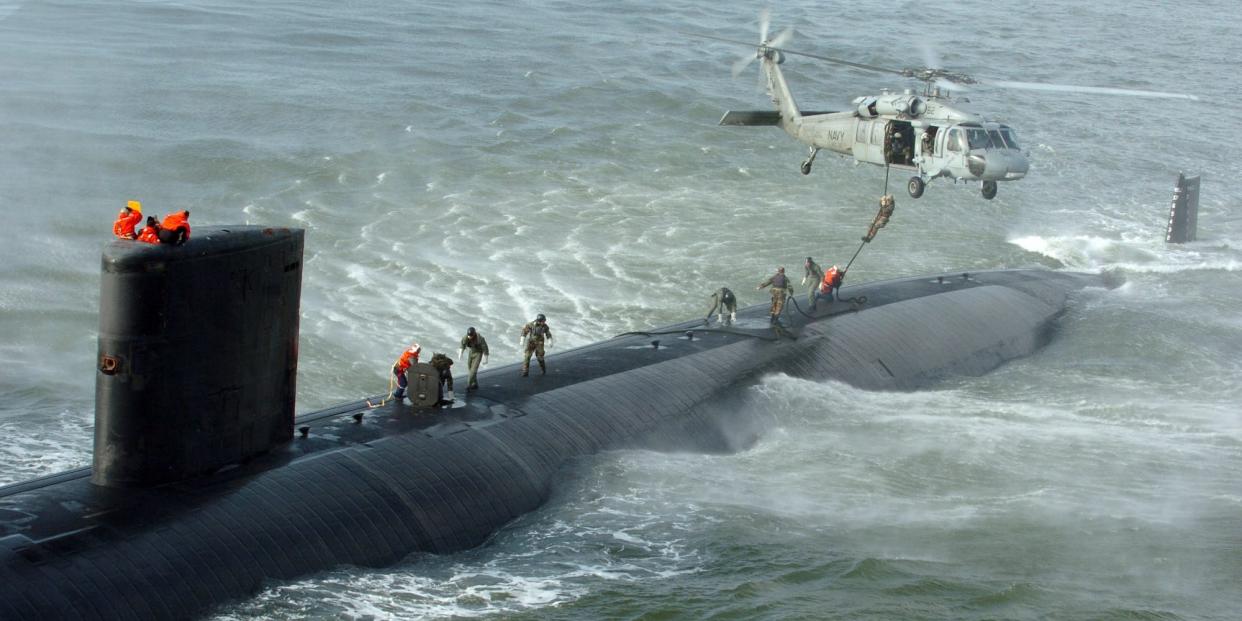 Navy SEALs run across the top of a submarine as a helicopter hovers overhead