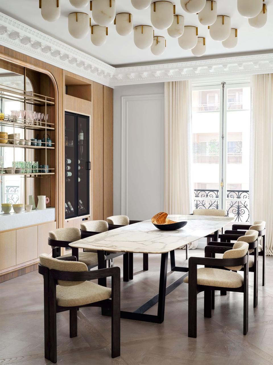 dining table with black base and marble top, eight black framed chairs with fabric seats, inset in wall with cabinets and open shelves with glassware, wine refrigerator, multiple alabaster ceiling lights, window