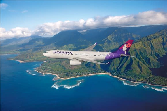 A Hawaiian Airlines plane flying over the ocean, with mountains in the background.