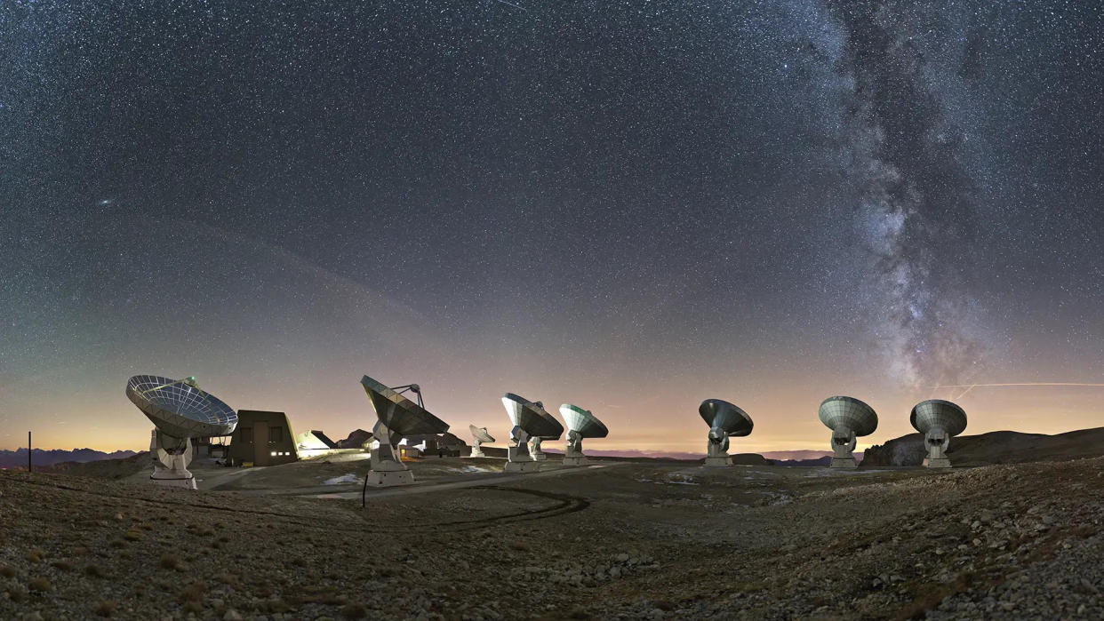  Several giant satellite dishes stand scattered in a vast rolling field, pointed toward the night sky and an array of stars next to the arm of the milky way. 