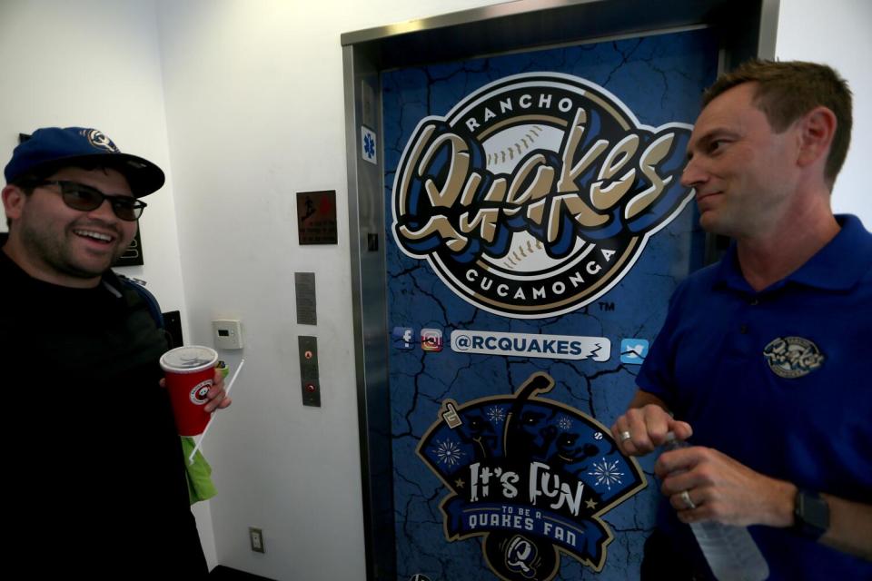 Two men talk in front of the logo for the Rancho Cucamonga Quakes