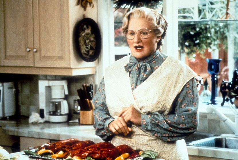 Robin William in a wig, glasses, an apron and grey dress standing in a kitchen for the movie "Mrs. Doubtfire"