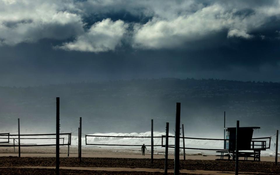 A man walks near volleyball courts as high surf pounds the shore.