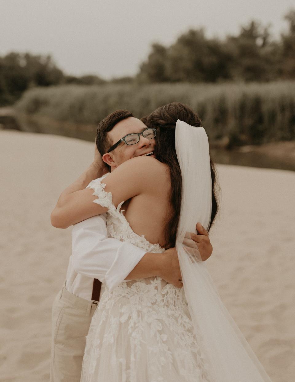 A brother hugs his sister, who wears a wedding dress, on a beach.