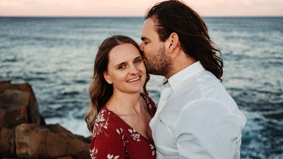 Michael gives Lisa a kiss on the cheek while she smiles at camera while at beach at sunset. 